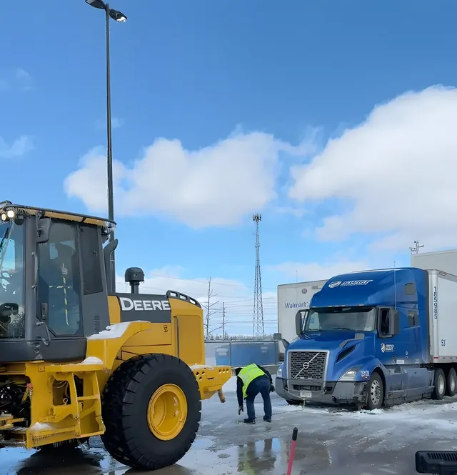 Yellow John Deere wheel loader parked on a wet lot beside a blue semi truck