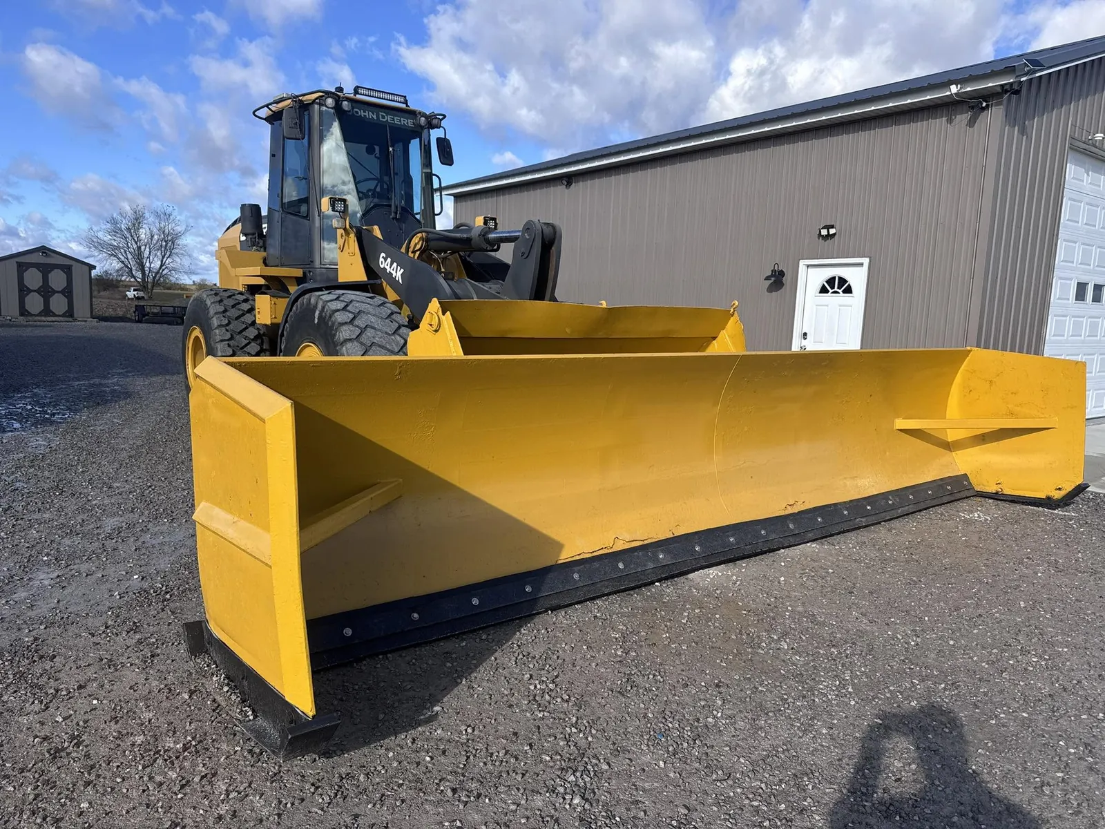 Yellow John Deere wheel loader with a long yellow pusher box parked on a gravel lot